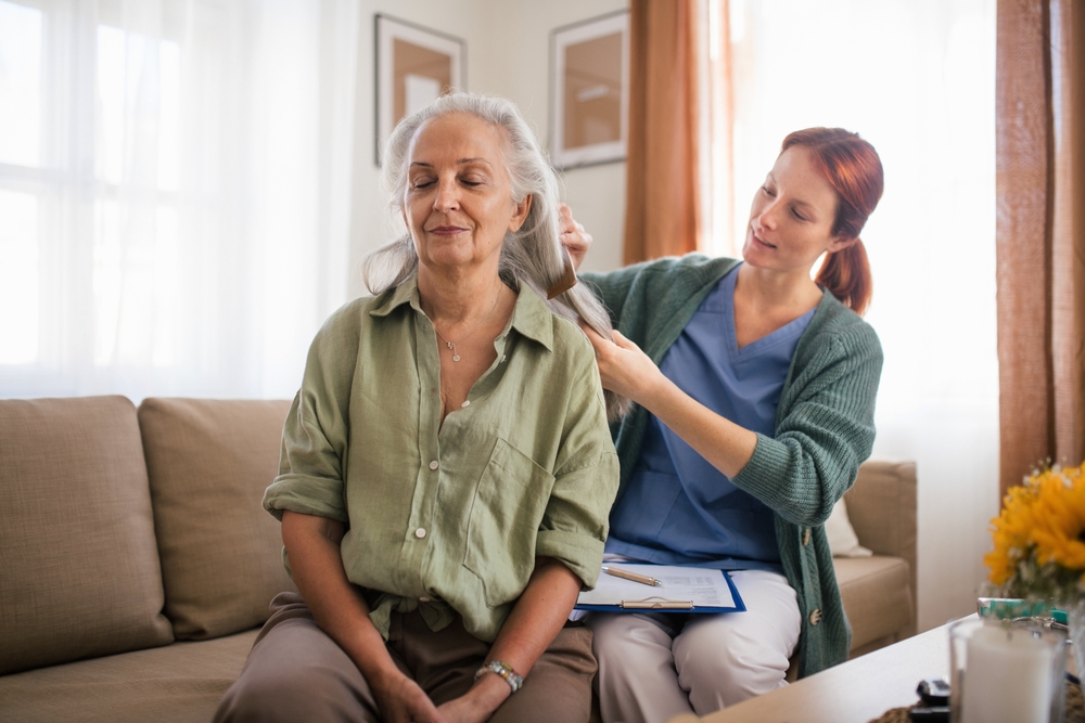 A caregiver brushing an elderly woman's hair as they sit on the couch at her home